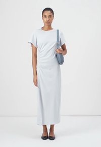 Svetlošedé maxi šaty s krátkymi rukávmi a bočnými nariaseniami; model drží malú modrú kabelku cez rameno a má na sebe čierne baleríny.