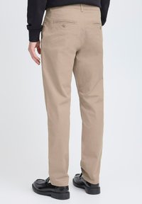 Pantaloni in cotone beige con vestibilità dritta, dotati di due tasche posteriori e scarpe nere a contrasto. Texture liscia, stile minimale.