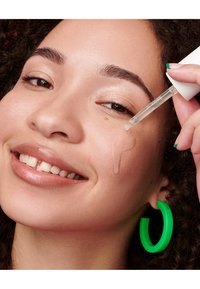 Klare serumdråper påført kinnet til en person med krøllete hår, som har grønne øredobber i form av ringer og en lys leppefarge.