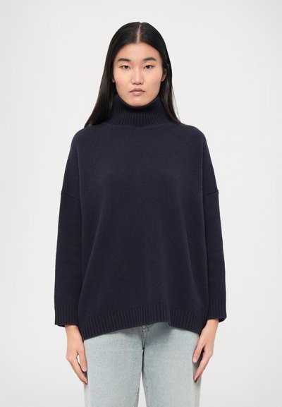 Mørkeblå rullekravesweater med løs pasform, ribbede manchetter og kant. Fremstillet af blødt, tekstureret materiale, båret over lyseblå jeans.