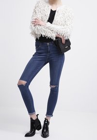 Vit texturerad fransad cardigan över en svart topp, kombinerad med ripped mörkblå skinny jeans och blanka svarta anklarstövlar. Svart handväska ingår.