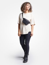 Borsa a tracolla nera indossata sopra una t-shirt oversize beige, abbinata a jeans skinny neri e stivali neri con lacci. Capelli ricci e sfondo neutro.