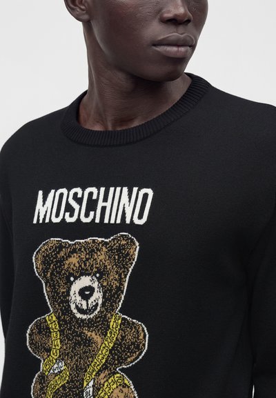 Gros plan sur une personne portant un pull noir avec un motif d'ours en peluche et le texte "MOSCHINO" au-dessus de l'ours.