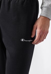 La mano destra poggiata accanto a dei pantaloni da tuta neri Champion con logo bianco sulla coscia, indossati con una maglietta grigia a maniche lunghe.