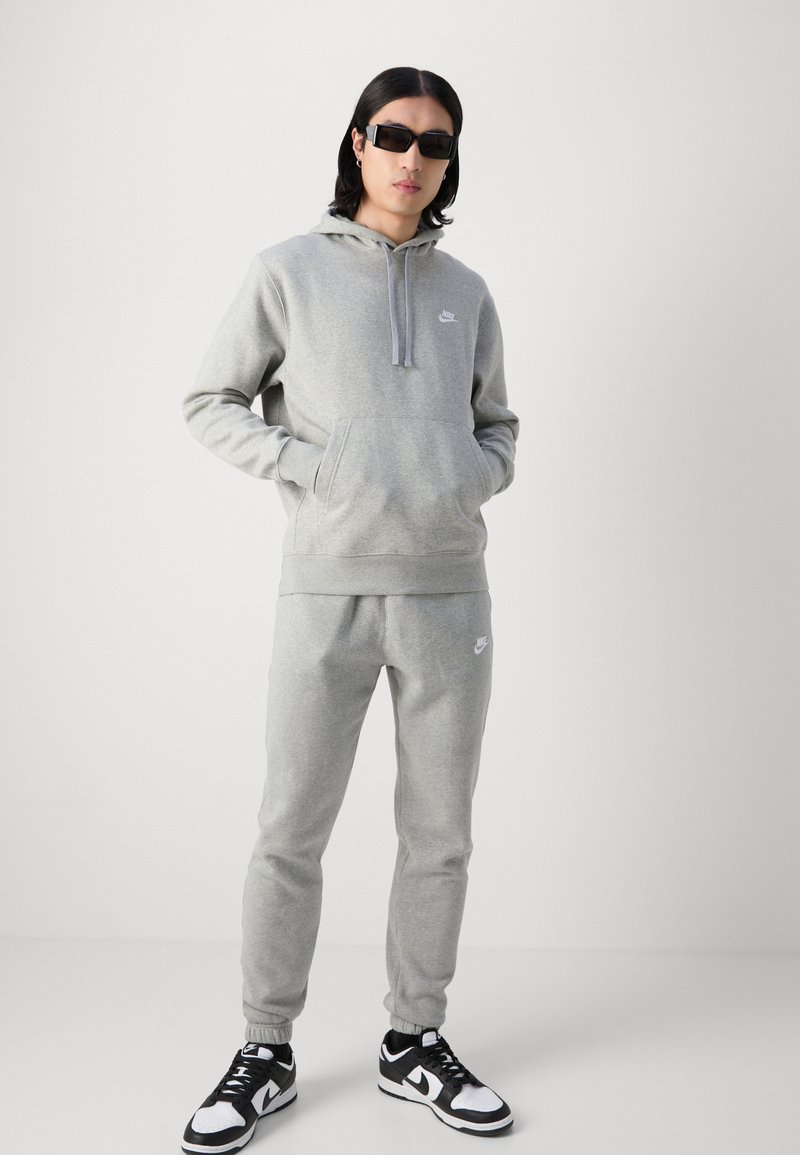 Graues Hoodie- und Hosen-Set aus weichem Stoff, mit einer Fronttasche und Logo-Akzenten. Kombiniert mit schwarz-weißen Sneakern.