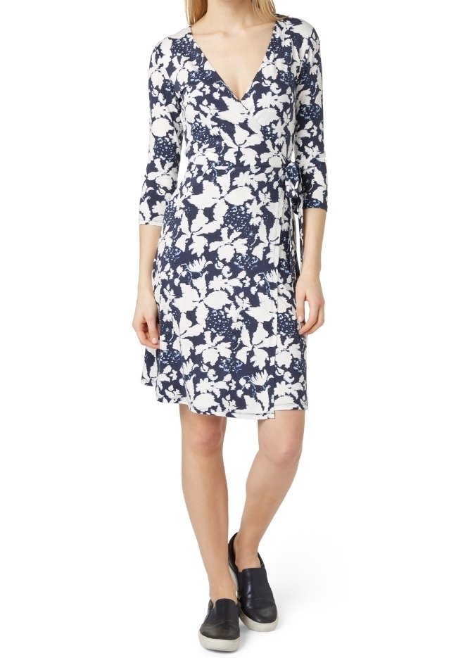 Femme portant une robe portefeuille fleurie bleu marine et blanche avec des manches trois-quarts et des chaussures noires sans lacets.