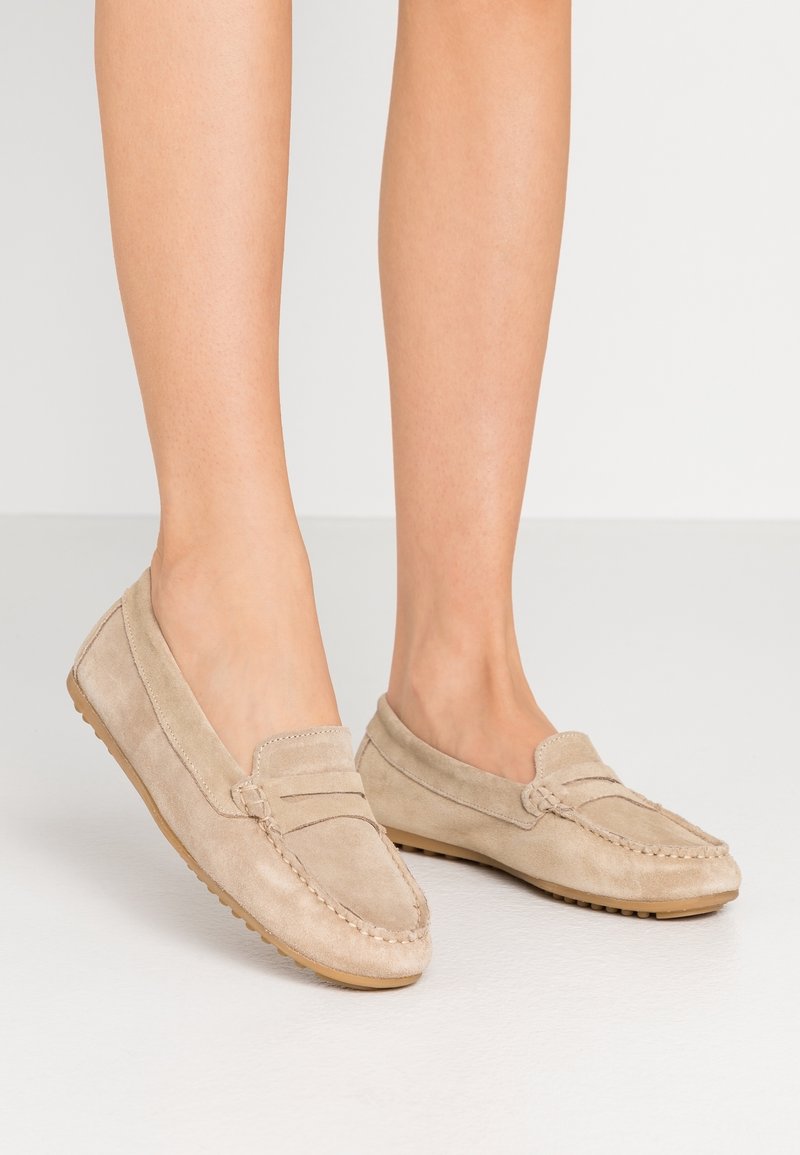 Pieds portant des mocassins en suède beige style mocassin avec des détails cousus et des semelles en caoutchouc sur un fond clair.