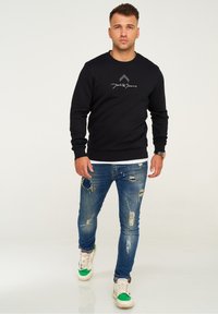 Black sweatshirt with a round neckline, featuring white brand logo. Worn with distressed blue jeans and green-accented sneakers.