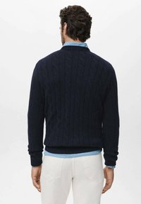 Suéter de punto de cable en color marino con dobladillo y puños acanalados, con cuello alto y llevado debajo de una camisa de mezclilla de color azul claro.
