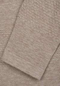 Gros plan sur un tissu de pull beige avec une surface texturée et douce et un poignet côtelé. Couleur claire avec un éclat subtil, mettant en valeur les variations de tissage.