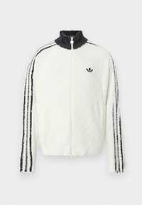 Chaqueta de felpa con cremallera en blanco, con cuello alto, logo de Adidas en negro y rayas negras a lo largo de las mangas. Textura suave y corte relajado.