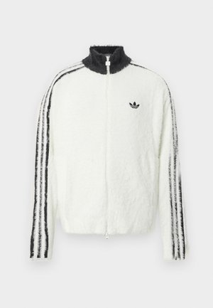 Apšiltintas zip-up džemperis baltos spalvos su aukštu apykakle, juodu Adidas logotipu ir juodomis juostelėmis palei rankoves. Minkšta tekstūra ir laisvas siluetas.