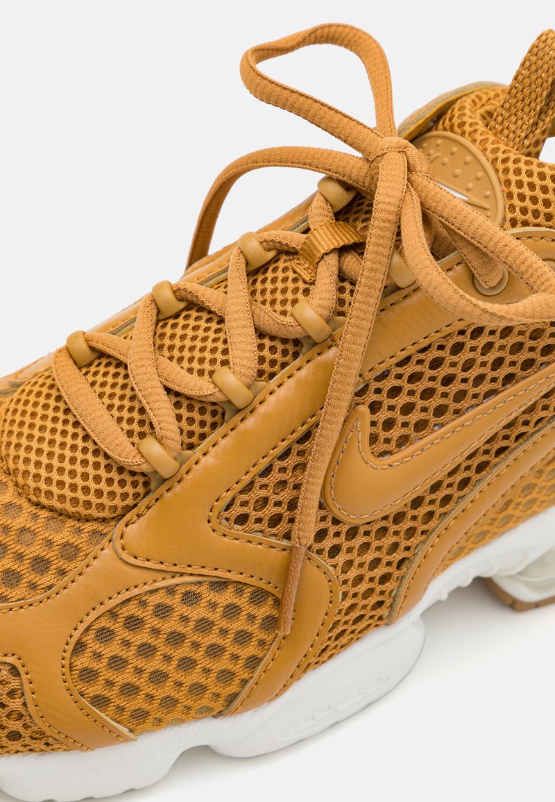 Baskets de sport beige avec un dessus en maille, des accents en cuir et des lacets ronds. Présente une semelle blanche et une surface texturée et respirante.