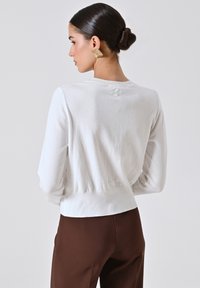 Donna con capelli scuri raccolti in uno chignon, che indossa orecchini quadrati dorati, una maglietta bianca a maniche lunghe e pantaloni marroni, vista di spalle su uno sfondo semplice.