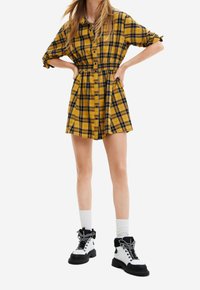 Donna che indossa un vestito a scacchi giallo e nero con maniche arrotolate, calze bianche e stivali allacciati in bianco e nero, in piedi con le mani sui fianchi.