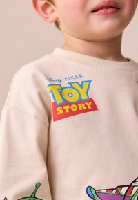Béžová bavlněná mikina s logem "Toy Story" v modré a červené barvě, s barevným grafickým motivem mimozemšťana na rukávu a žebrovaným výstřihem.