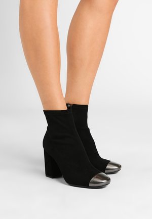Jambes portant des bottines en daim noir avec des bouts arrondis métalliques argentés et des talons hauts épais sur un fond gris clair.