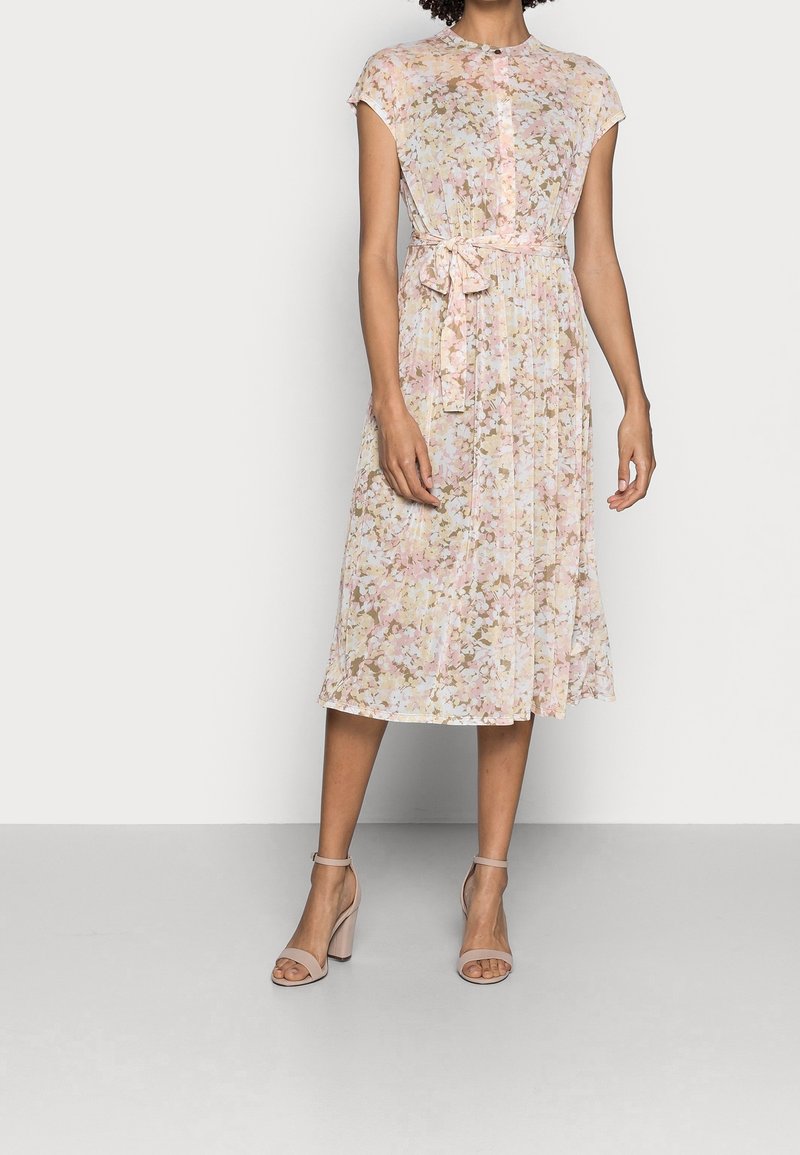 Femme portant une robe midi rose à fleurs avec manches courtes et ceinture nouée à la taille, associée à des talons blocs nude avec bride à la cheville.