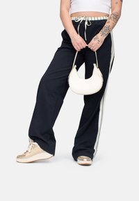Dunkelblaue weite Hose mit Kordelzugbund und weißen Seitenstreifen. Kombiniert mit metallisch goldenen Sneakern und einer crèmefarbenen Handtasche.