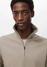 Suéter ligero beige claro con media cremallera y un tejido texturizado. El cuello es alto, creando un aspecto deportivo.