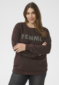 Hnedý sveter s nápisom "FEMME" v trblietkom písme. Obsahuje okrúhly výstrih, dlhé rukávy a rebrované manžety. Skombinované s tmavými džínami.