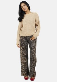 Pull en maille beige à coupe loose, associé à un pantalon large à imprimé léopard noir et marron. Le modèle porte des chaussures rouges.