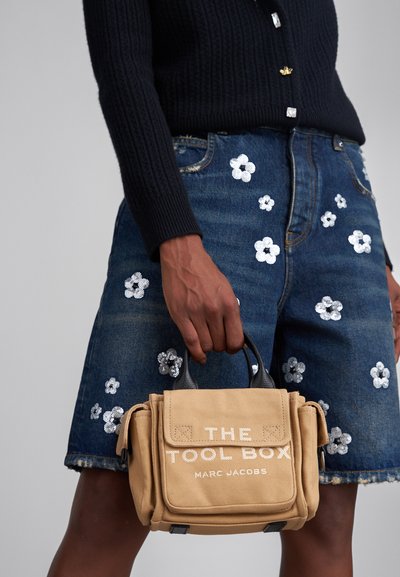Beige canvas handtas met bovenhandvaten, met zwarte accenten en witte tekst waarop "THE TOOL BOX" en "MARC JACOBS" staat. Denimrok met bloemenversieringen.