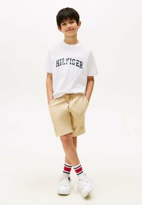 Hvid T-shirt med marineblå "HILFIGER" print, beige shorts og hvide sneakers. Sokker med røde, hvide og blå striber. Afslappet outfit.