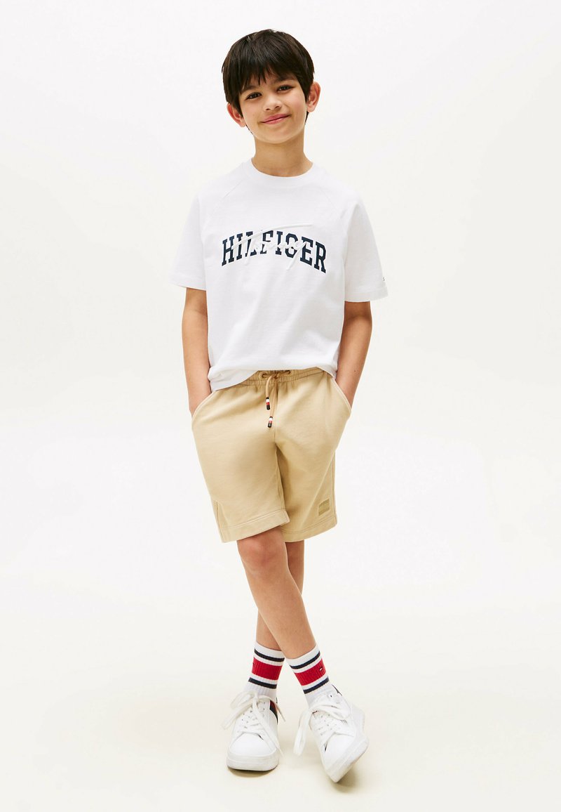 Hvid T-shirt med marineblå "HILFIGER" print, beige shorts og hvide sneakers. Sokker med røde, hvide og blå striber. Afslappet outfit.