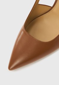 Chaussure en cuir marron à bout pointu avec un design découpé sur le côté, présentant une texture lisse et un intérieur minimaliste.