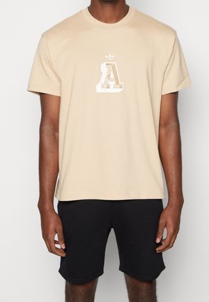 Beige katoenen T-shirt met een witte grafische "A" logo op de borst. Combinatie met zwarte shorts, wat een casual pasvorm laat zien.