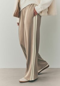 Un pantalon de survêtement beige présente une coupe décontractée, des poches latérales et des rayures verticales blanches le long des coutures extérieures, associé à des chaussures de couleur claire.