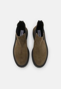 Hnědé semišové slip-on boty s hladkým povrchem, elastickými bočními panely a černou gumovou podrážkou. Minimalistický design bez viditelného hardwaru.