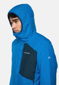 Blauwe geïsoleerde jas met een capuchon, voorzien van een donkergroene zijpaneel, rits aan de voorkant en het Vaude-logo op de borst. Soepele textuur.
