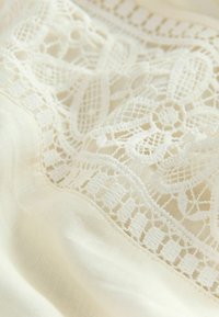 Țesătură de dantelă într-o nuanță delicată de fildeș, cu modele florale complicate și o textură fină, ușoară, prezentând cusături delicate de-a lungul marginii.