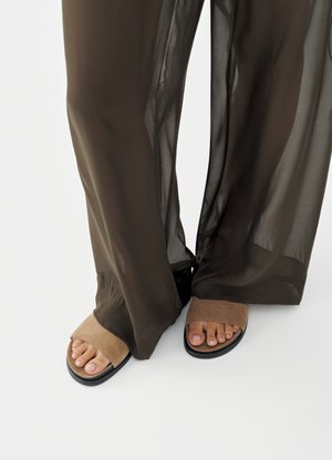 Πόδια με ταμπά σουέντ σανδάλια ανοιχτής μύτης, μερικώς καλυμμένα από διάφανο σκούρο καφέ παντελόνι με φαρδιά πόδια, σε λευκό φόντο.
