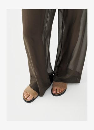 Picioare în sandale deschise la degete din piele întoarsă de culoare bej, parțial acoperite de pantaloni largi maro închis, transparenți, pe fundal alb.