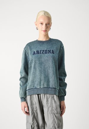 Blue sweatshirt made of soft fabric, featuring "ARIZONA" in navy letters across the chest and ribbed cuffs and hem. Paired with gray cargo pants.