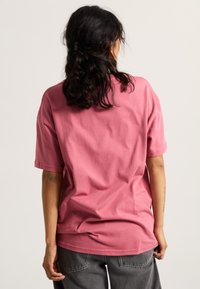 Camiseta de algodón rosa con mangas cortas, corte relajado y cuello redondo, vista desde la parte posterior. Pantalones grises visibles por debajo.