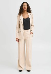 Beige taillierter Blazer und weite Hose kombiniert mit einem schwarzen Camisole mit Spitzenbesatz. Glattes Material mit strukturierter Silhouette. Schwarze spitze Schuhe.