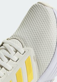 Scarpa da ginnastica grigia chiara con tomaia testurizzata, lacci bianchi e un accento giallo di lato. Presenta un colletto imbottito e una suola ammortizzata.