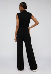 Czarny bezrękawnikowy kombinezon z wysokim kołnierzem, szerokimi nogawkami i prążkowanymi detalami. Wykonany z gładkiego materiału, posiada dopasowaną talię i luźny sylwetkę.