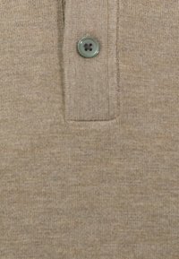 Polo shirt color sabbia con tessuto texturizzato, colletto classico e un singolo bottone verde con un dettaglio centrale nero.