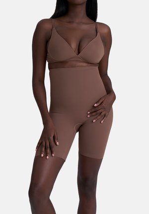 Vrouw in bruine hooggewaardeerde shaping shorts en bijpassende beha, staande met één hand op haar bovenbeen en de andere op haar heup tegen eenwitte achtergrond.