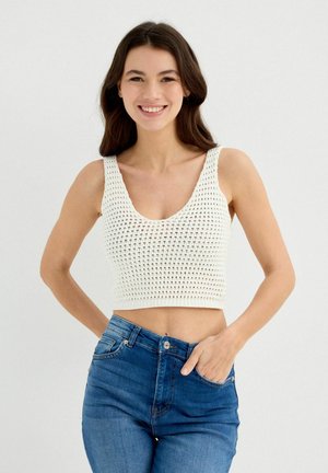 Top corto de crochet blanco con tirantes anchos, escote profundo y diseño texturizado, combinado con jeans de mezclilla azul de talle alto.