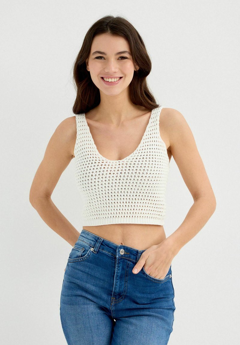 Top corto de crochet blanco con tirantes anchos, escote profundo y diseño texturizado, combinado con jeans de mezclilla azul de talle alto.