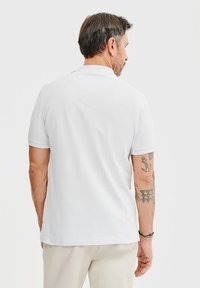 Biała koszulka polo z klasycznym kołnierzykiem i krótkimi rękawami, z teksturowanej tkaniny. Widok z tyłu ukazuje tatuaże na ramieniu noszącego.