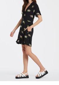 Black dress with short sleeves and a notched collar, featuring floral patterns, two side pockets, and button detailing. Paired with white strappy sandals.