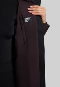 Abrigo negro y burdeos con diseño acolchado, que muestra un parche de marca visible en el interior. Presenta una textura suave y una forma ajustada.