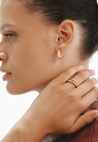 Pendientes de oro con un diseño liso y minimalista, y un anillo texturizado a juego en una mano que descansa sobre un hombro.